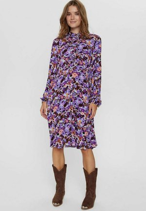 Blumenkleid in lila Tönen mit langen Ärmeln, durchgehender Knopfleiste und elastischen Bündchen. Kombiniert mit braunen Cowboystiefeln.