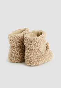 Botines beige hechos de un tejido texturizado y rizado con un interior esponjoso. Presentan un puño doblado y una forma redondeada, proporcionando una cómoda cobertura para los pies.
