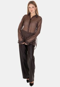 Blusa marrone trasparente con pois bianchi, chiusura frontale con bottoni, maniche lunghe. Abbinata a pantaloni neri lucidi a vita alta e tacchi neri.