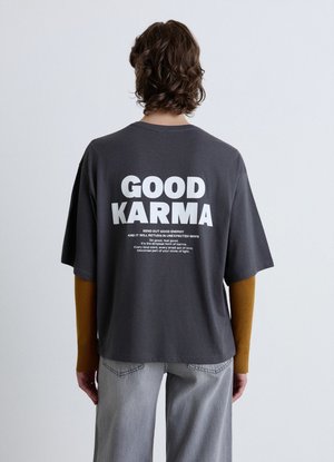 Persona con capelli ricci che indossa una maglietta oversize grigio scuro con testo bianco "GOOD KARMA" e testo motivazionale più piccolo sul retro.