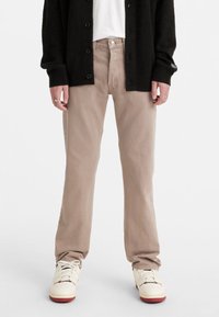Jeans rectos de color beige claro, confeccionados en algodón, con bolsillos frontales y cierre de botones, combinados con zapatillas blancas y un cárdigan negro.