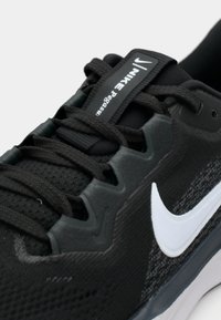 Pantof sportiv negru cu partea superioară din plasă texturată, logo-ul Nike alb și șireturi negre. Dispune de o talpă căptușită și de un sistem de sprijin întărit.