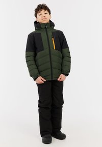 Groene en zwarte gewatteerde jas met capuchon, voorzien van een oranje ritszak en textuurpanelen, gedragen met zwarte broeken en laarzen.