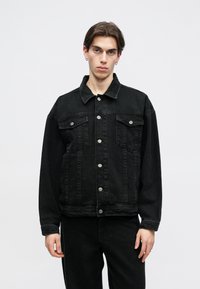 Jaqueta de ganga preta com fecho de botões à frente, dois bolsos ao peito e colarinho clássico. Apresenta um design ligeiramente ajustado e textura resistente.