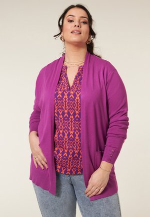 Paarse cardigan met open voorkant, geribbelde manchetten en zakken over een v-hals top met een patroon in paars en oranje. Draagt bij lichte blauwe spijkerbroek.