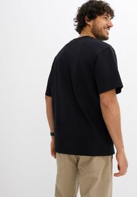 Un uomo con capelli ricci sorride oltre la spalla indossando una camicia nera a maniche corte e pantaloni beige su uno sfondo bianco semplice.