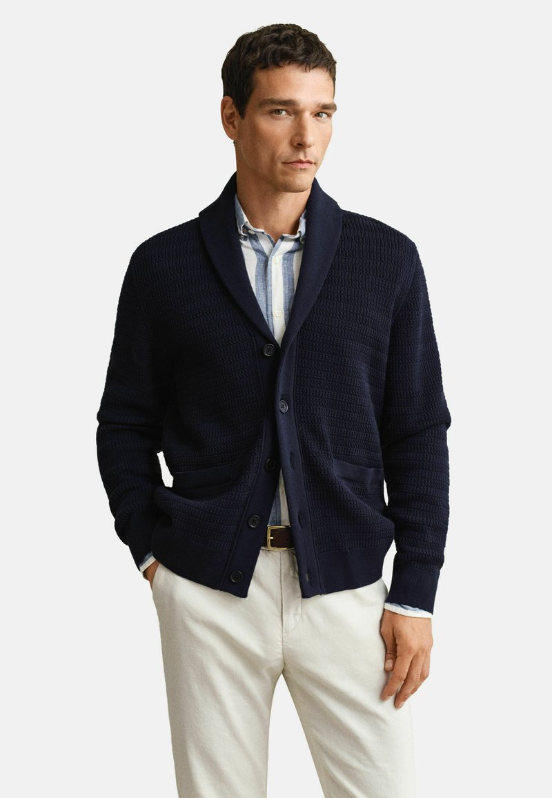 Homme portant un cardigan boutonné texturé bleu marine sur une chemise rayée bleu clair avec un pantalon beige et une ceinture marron.