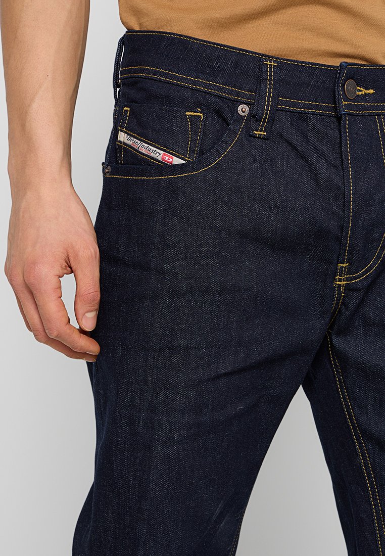Dunkelblaue Jeans aus Denim mit gelben Nähten, einer Vorderasche, die ein Etikett zeigt, und einer standardmäßigen Vorderreißverschluss- und Knopfverschluss.