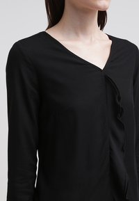 Czarny longsleeve z dekoltem w serek i falbanami z przodu. Wykonany z miękkiego, gładkiego materiału, charakteryzuje się prostym wzornictwem.