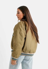 Olijfgroene bomberjack met ribgebreide manchetten en tailleband, voorzien van blauwe en witte strepen. Draagt over lichtblauwe jeans, zijaanzicht weergegeven.