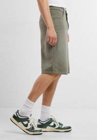 Pantaloni corti verde oliva in cotone, con un taglio classico, tasche laterali e orli cuciti, abbinati a sneakers verdi e bianche.
