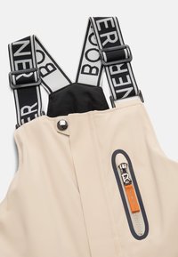 Béžové kšandy s černobílými nastavitelnými elastickými popruhy s výrazným textem a zipovou kapsou na hrudi s oranžovým táhlem.