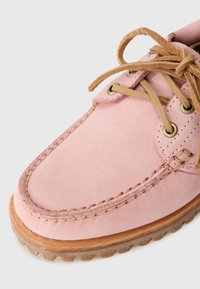 Close-up of a pink suede shoe with beige laces, detailed stitching, brass eyelets, and a textured brown rubber sole.