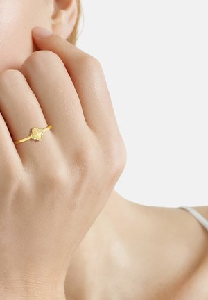 Mano con anillo dorado en forma de flor tocando el rostro cerca de la oreja sobre un fondo claro y liso.