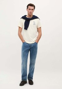 Crème t-shirt met blauw logo, gecombineerd met lichtblauwe spijkerbroek. Een marineblauwe trui over de schouders gedrapeerd, zwarte schoenen met veters.