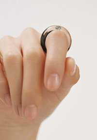 Anillo negro y plateado en la punta de un dedo, con un acabado suave y una pequeña piedra preciosa transparente. La banda es redondeada y tiene un color contrastante.