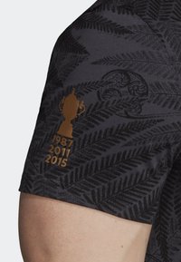 Mörkgrå t-shirt med ett ormbunksmönster och en guldtrophédesign på ärmen, tillsammans med åren 1987, 2011 och 2015.