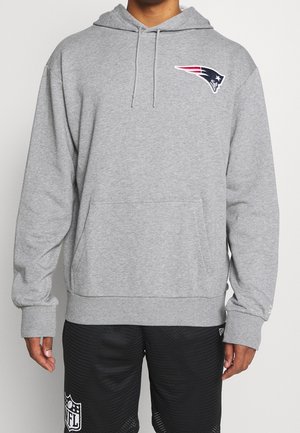 Person iført grå hættetrøje med New England Patriots-logo og sorte NFL-shorts står mod ensfarvet baggrund.