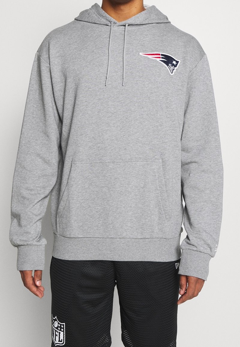 Personne portant un sweat à capuche gris avec le logo des New England Patriots et un short NFL noir, debout devant un fond uni.
