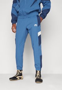 Blauwe trainingsbroek met elastische tailleband, zijzakken en witte en marineblauwe ontwerpelementen. Gemaakt van lichtgewicht stof met een gladde textuur.