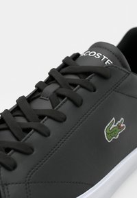 Fekete bőr sneaker lapos cipőfűzővel, fehér gumitalppal, zöld hímzett Lacoste logó piros kiegészítéssel, sima felületi textúrával.