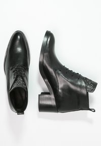 Botas de tobillo de cuero negro con puntera redonda, cordones en la parte delantera y ojetes metálicos decorativos en el caña. Tacón grueso y apilado.