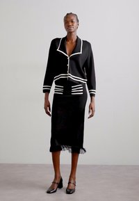 Cardigan negru din tricot cu dungi albe, cu închidere cu nasturi în față și fustă midi asortată cu un tiv franjurat. Purtat cu pantofi cu toc negri.