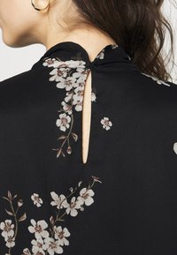 Haut en tissu noir présentant des motifs floraux en blanc et beige, avec une ouverture en forme de goutte d'eau au dos et un col rond.