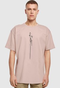 Camiseta de manga corta de color rosa claro, hecha de algodón. Presenta un diseño gráfico negro con la palabra "fe" alineada verticalmente en el centro.