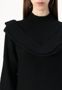 Juoda žakardo megzta kedro su aukštu apykakle ir plačiu, banguotu kaklo iškirpimu. Audinys yra minkštas ir storas, pabrėžiantis šilumą ir komfortą.