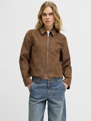 Frau mit hellbraunen Haaren trägt braune Wildlederjacke mit Reißverschluss und hellblaue Jeans, die Hände in den Taschen, vor einfarbigem Hintergrund.