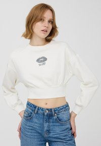 Biała, krótka bluza z długimi rękawami, z nadrukiem srebrnych ust oraz napisem "Więcej miłości, proszę", zestawiona z jeansami o wysokim stanie.