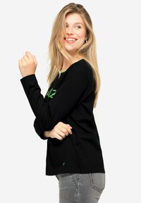 Zwarte langslavige shirt met groene grafische tekst en een ronde halslijn. Gemaakt van zacht materiaal, met een relaxte pasvorm en een subtiel logodetail.