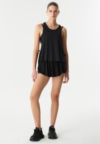 Top sin mangas atlético negro de ajuste holgado, diseño en capas y lazos laterales ajustables, combinado con pantalones cortos negros y zapatos deportivos.