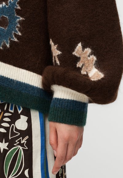 Κοντινό πλάνο ενός χεριού που φοράει καφέ μάλλινο πουλόβερ με λουλουδένια σχέδια και ριμπ μανσέτες πάνω από μια φούστα με σχέδια αφηρημένων σχημάτων.