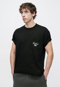 Μαύρο βαμβακερό t-shirt με κοντά μανίκια, διαθέτει τσέπη στο στήθος με λευκό κεντημένο λογότυπο. Ομαλή υφή, άνετη εφαρμογή.