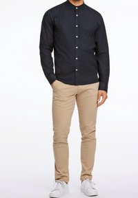 Camisa negra de botones de manga larga con cuello mao; combinada con pantalones beige y zapatillas blancas, creando un atuendo casual.