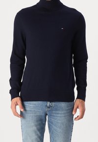 Marinblå polo-tröja i mjukt material med långa ärmar och ribbade muddar. Litet logotypdetalj på bröstet. Kombinerad med ljusblå jeans.