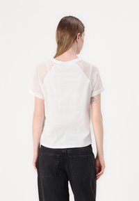 T-shirt bianco con maniche raglan trasparenti e scollatura arrotondata, abbinato a jeans neri a vita alta. Sono visibili dettagli di tessuto testurizzato.