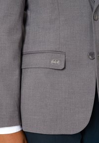 Grauer Blazer mit glatter Textur, der einen Satinkontur am Ärmel und ein kleines Metall-Logo-Akzent auf der Tasche aufweist.