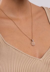 Eine roségoldene Halskette mit einem runden Anhänger und einem kleinen klaren Akzentstein, die an einer feinen Kette gegen ein neutrales, geripptes Oberteil hängt.