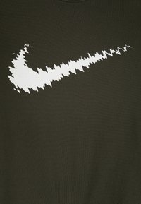 Oliwkowa koszulka z dużym białym logo Nike w kształcie „swoosh”, lekko teksturowana, z poszarpanym wzorem, wykonana z miękkiego materiału.