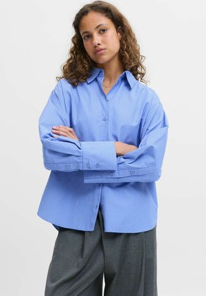 Chemise à carreaux bleus, à manches longues, avec une coupe décontractée et de larges poignets. Le tissu semble lisse avec une texture subtile.