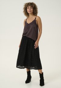 Camisole en dentelle marron foncé associée à une longue jupe noire brodée, ornée de motifs floraux et d'une silhouette évasée, portée avec des bottines noires.