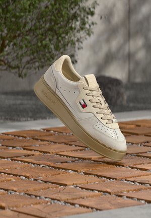 Beżowy i biały sportowy but na co dzień z beżową podeszwą i sznurowadłami, z małym logo w kolorach czerwonym, białym i niebieskim po boku, unoszący się nad teksturowaną podłogą.