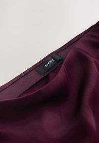 Gros plan sur un tissu violet foncé avec une étiquette de vêtement noire cousue indiquant "NEXT" sur une ceinture élastique.