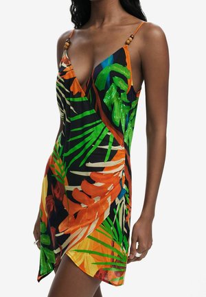 Vrouw draagt een mouwloze wikkeljurk met V-hals met een oranje, groene, blauwe en zwarte tropische bladprint en dunne bandjes met houten kraaldetails.