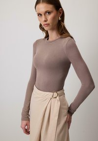 Woman wearing a fitted long-sleeve mauve top and a cream wrap skirt with a gold buckle, standing against a plain light background.