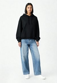 Svart bomullshoodie med ficka fram och dragsnören, tillsammans med lösa blå denimbyxor. Modellen står på en ljus bakgrund.