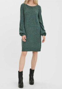 Robe longue à manches longues en tricot vert avec un col bateau, présentant une coupe décontractée et des poignets côtelés. Portée avec des bottines à plateforme noires.